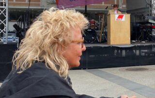 Beate Müller-Gemmeke bei einer 1.-Mai-Kundgebung des DGB. Im Vordergrund im Profil, im Hintergrund eine Rednerin am DGB-Rednerpult auf einer Bühne.