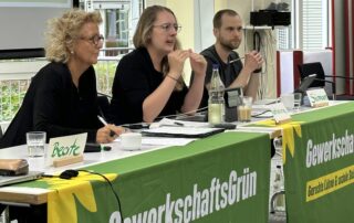 eate Müller-Gemmeke bei einer Sitzung von GewerkschaftsGrün. Sie sitzt neben anderen Teilnehmenden hinter einem Tisch mit grünem Banner und diskutiert konzentriert.