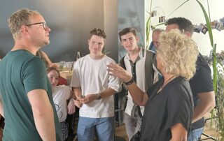 Beate im lebendigen Gespräch mit einer Gruppe junger Menschen – darunter auch eine Person im Rollstuhl. Im Hintergrund sind Deko, Getränke und die Fotowand sichtbar.