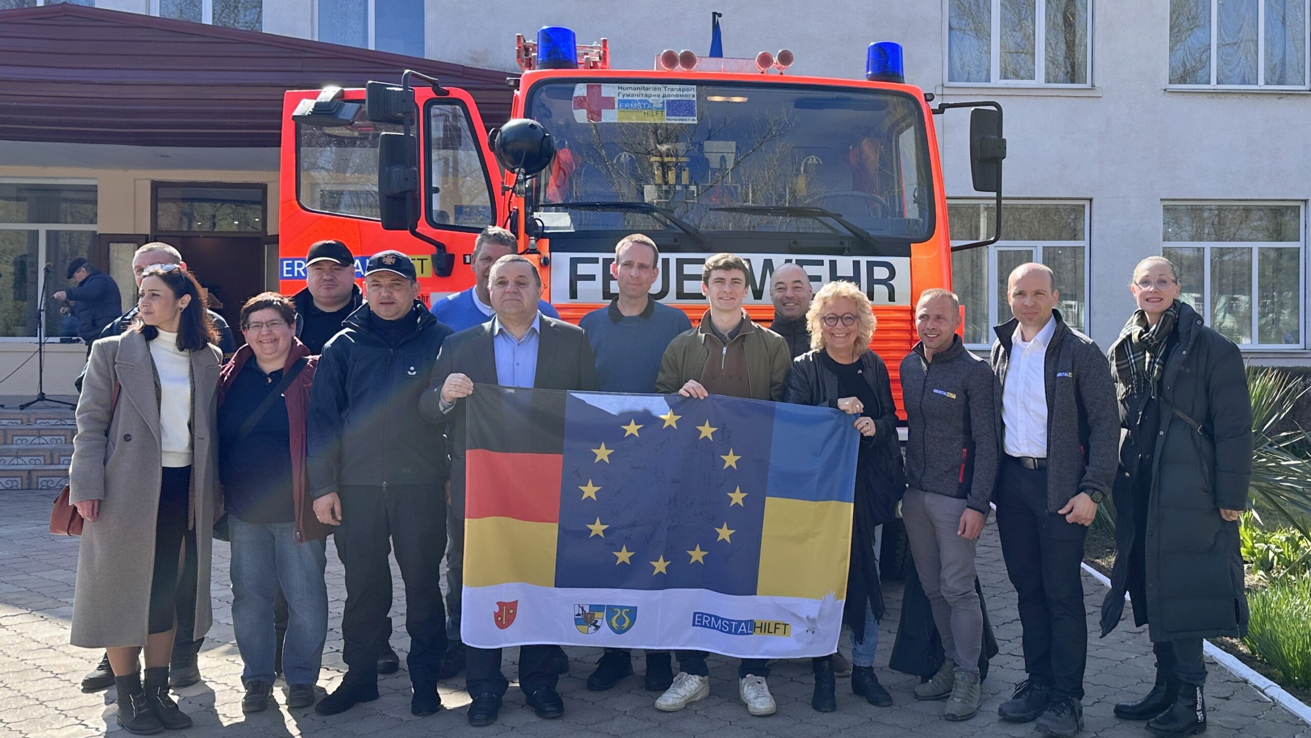 Gruppenfoto vor einem roten Löschfahrzeug bei der Übergabe in Bessarabske. In der Mitte: Beate Müller-Gemmeke. Die Flagge vereint EU, Deutschland und Ukraine.