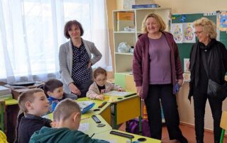 Besuch in einem Klassenzimmer in Arzis in der Ukraine: Kinder sitzen an grünen Tischen, zwei Lehrerinnen und Beate Müller-Gemmeke stehen daneben und beobachten den Unterricht.