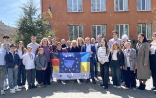Gruppenbild vor einem Schulgebäude in Arzis: Die Delegation aus Deutschland und die ukrainischen Gastgeber:innen halten eine Solidaritäts-Flagge mit EU-, Ukraine- und Deutschlandfahne.