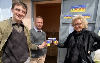 Drei Mitglieder der Delegation stoßen mit Trinkbechern an – im Hintergrund die geöffnete Tür zur Trinkwasseraufbereitungsstation, unterstützt durch die Solidaritätspartnerschaft.