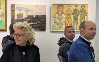 Beate Müller-Gemmeke sitzt mit anderen Delegierten in einer Galerie, umgeben von Kunstwerken, darunter ein Bild mit Soldaten und ein traditionelles Motiv.