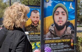 Beate Müller-Gemmeke steht vor Gedenktafeln mit Porträts gefallener Soldaten. Sie blickt auf das Bild eines jungen Mannes, umgeben von Blumen und Fahnen in Blau und Gelb.