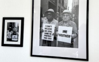 Zwei gerahmte Schwarz-Weiß-Fotos an einer weißen Wand. Das große Bild zeigt zwei ältere Frauen bei einer Demonstration mit Schildern „I can’t believe we still have to protest this shit“ und „Me neither“. Im kleineren Bild wurde Beate Müller-Gemmeke digital als dritte Person eingefügt – mit einem eigenen Schild „Me neither“.
