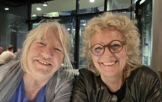 Beate Müller-Gemmeke und Hanne nebeneinander an einem Tisch, beide lachen herzlich in die Kamera, Hanne trägt ein blaues Oberteil, Beate eine schwarze Lederjacke.