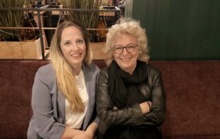 Beate Müller-Gemmeke und Janina sitzen nebeneinander auf einem Sofa, beide lächeln, Janina trägt ein hellblaues Blazer-Jäckchen, Beate ist ganz in Schwarz gekleidet.