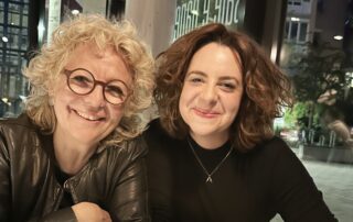 Beate Müller-Gemmeke und Lisa am Tisch, beide blicken freundlich in die Kamera, Lisa trägt ein schwarzes Oberteil, Beate eine Lederjacke und Brille, im Hintergrund große Fenster.