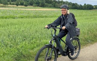 Beate Müller-Gemmeke fährt mit dem E-Bike auf einem Feldweg über die Schwäbische Alb – im Hintergrund grüne Felder und sanfte Hügel. Sie trägt Helm, Brille und wetterfeste Kleidung und lächelt in die Kamera.