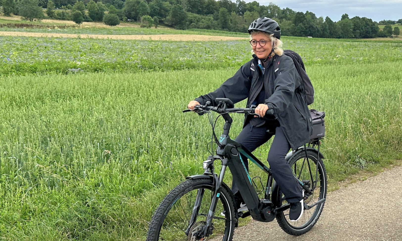 Beate Müller-Gemmeke fährt mit dem E-Bike auf einem Feldweg über die Schwäbische Alb – im Hintergrund grüne Felder und sanfte Hügel. Sie trägt Helm, Brille und wetterfeste Kleidung und lächelt in die Kamera.