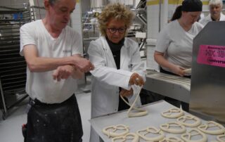 Beate Müller-Gemmeke formt unter Anleitung eine Brezel bei BeckaBeck – beide in Bäckerjacken, konzentriert am Teigtisch.