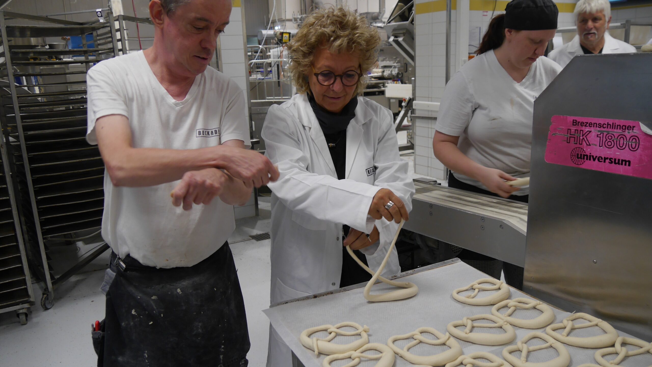 Beate Müller-Gemmeke formt unter Anleitung eine Brezel bei BeckaBeck – beide in Bäckerjacken, konzentriert am Teigtisch.