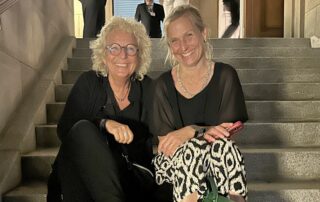 Beate Müller-Gemmeke sitzt auf einer Steintreppe gemeinsam mit Katja Keul (ehemalige grüne MdB). Beide lachen, es ist Abendstimmung beim Sommerfest der Deutschen Parlamentarischen Gesellschaft.