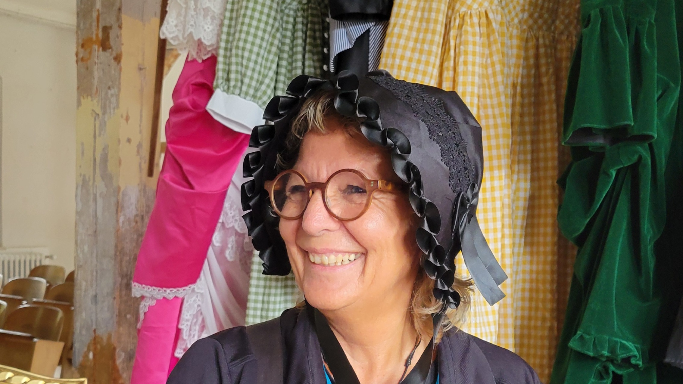 Beate Müller-Gemmeke mit schwarzer historischer Haube vor bunten Kleidern – fröhlich und verkleidet beim Rundgang durchs Albgut.