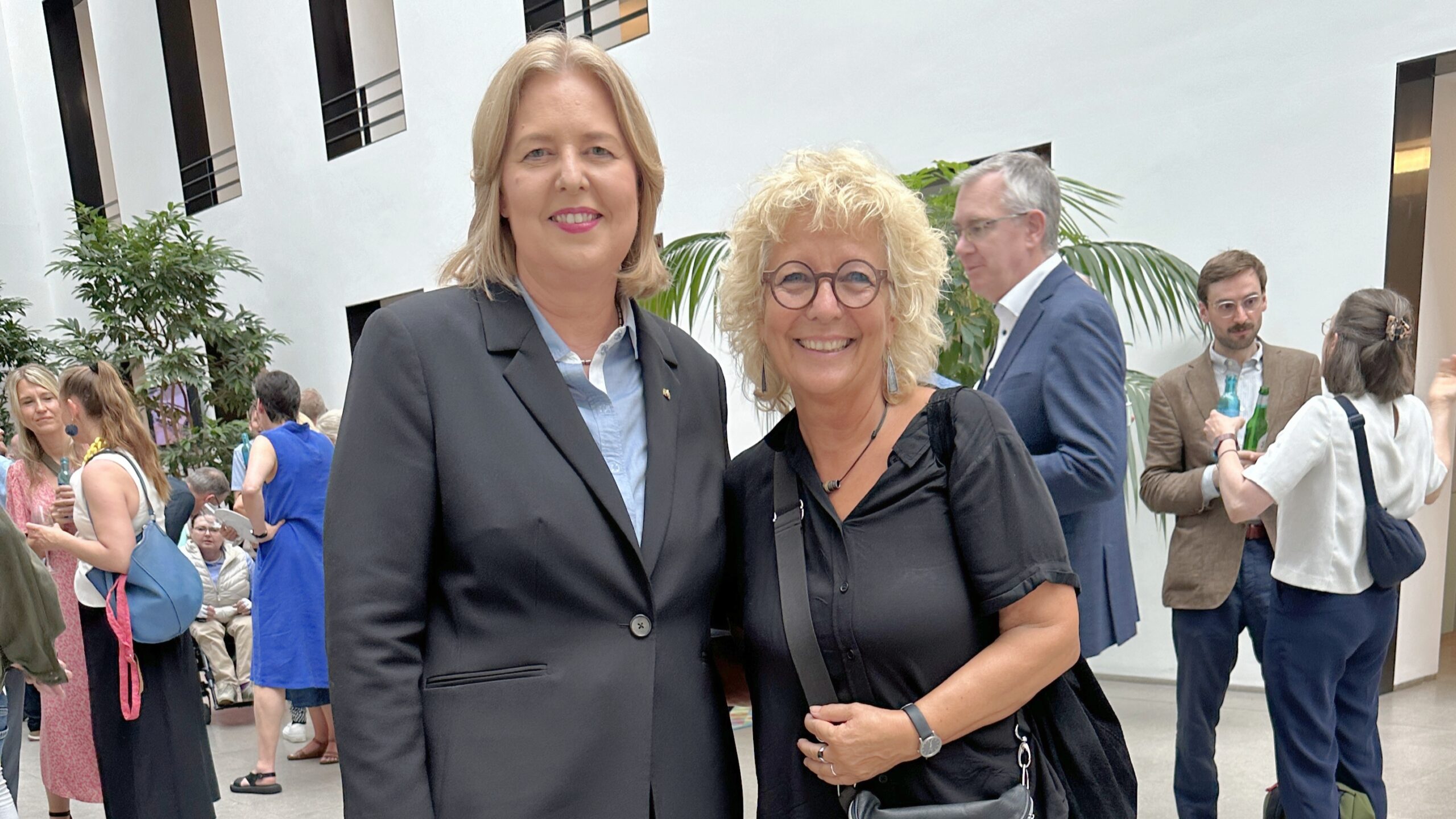 Beate Müller-Gemmeke zusammen mit Bärbel Bas, der neuen Bundesministerin für Arbeit und Soziales, beim Hoffest des BMAS. Beide stehen in einem modernen Innenhof und lächeln in die Kamera.
