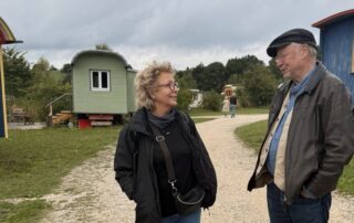 Beate Müller-Gemmeke im Gespräch mit einem Mann auf dem Gelände der Hopfenburg – bunte Zirkuswagen säumen den Weg, der Himmel ist grau.