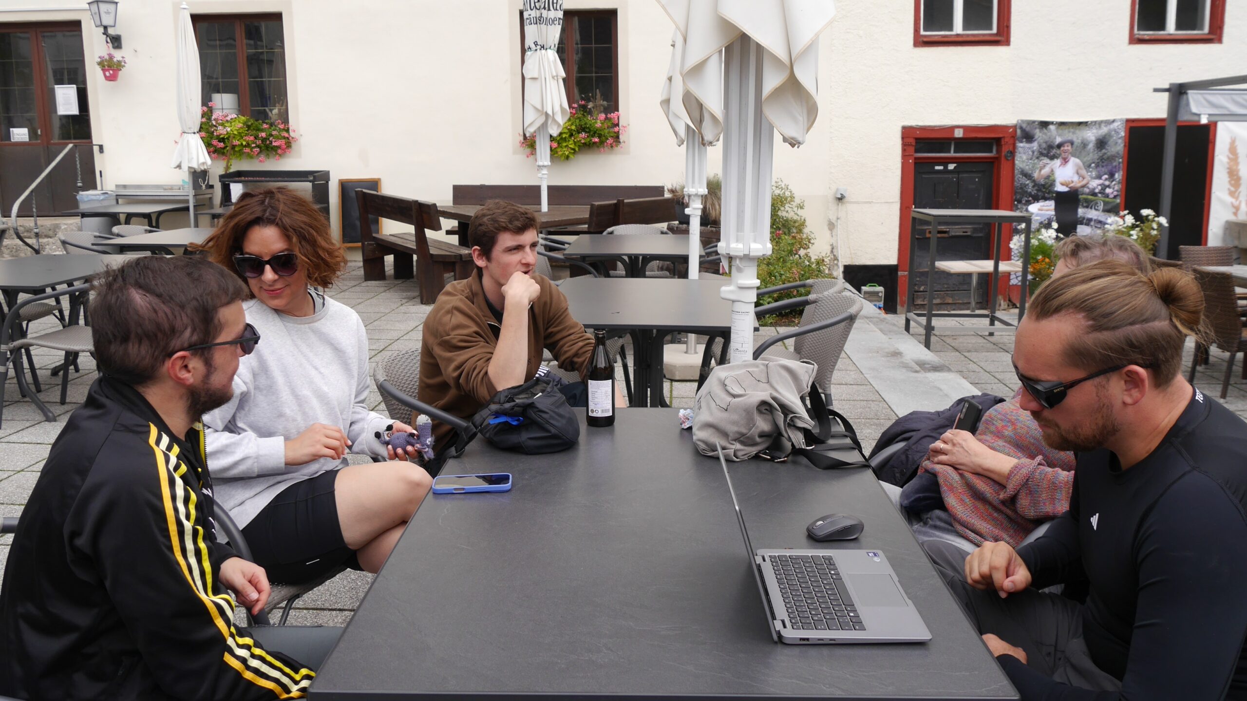 Fünf Personen – darunter Niklas, Lisa, Jaron und Christian – sitzen an einem großen Tisch im Außenbereich eines Cafés. Ein Laptop steht auf dem Tisch, einige unterhalten sich, andere wirken konzentriert. Die Stimmung ist ruhig, es ist eine Pause während der Albtour.