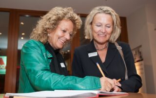 Beate Müller-Gemmeke und Sylvia Kotting-Uhl tragen sich gemeinsam in ein Gästebuch ein und lachen dabei herzlich.