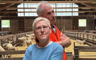 Simone und Christian stehen Rücken an Rücken in einem offenen Schafstall. Im Hintergrund sind mehrere Schafe in abgetrennten Bereichen zu sehen. Beide blicken ernst, aber mit einem Augenzwinkern in die Kamera.