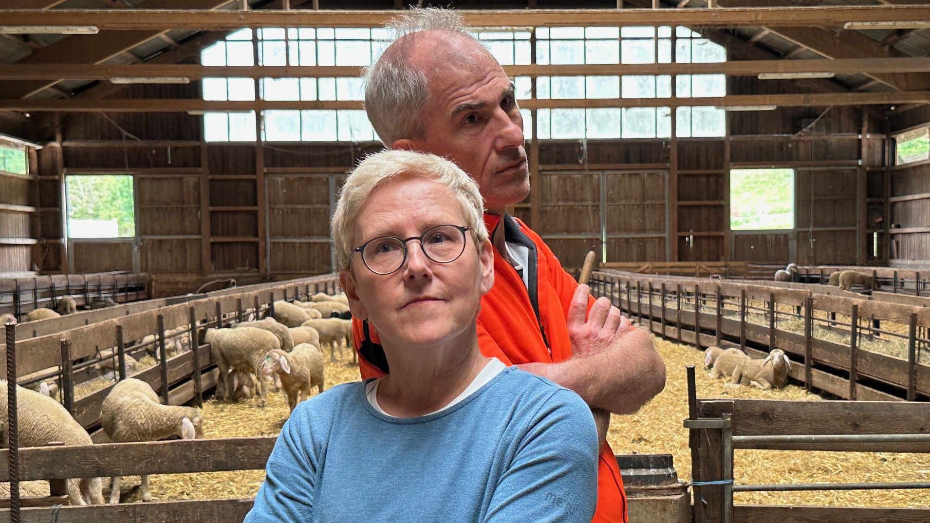 Simone und Christian stehen Rücken an Rücken in einem offenen Schafstall. Im Hintergrund sind mehrere Schafe in abgetrennten Bereichen zu sehen. Beide blicken ernst, aber mit einem Augenzwinkern in die Kamera.