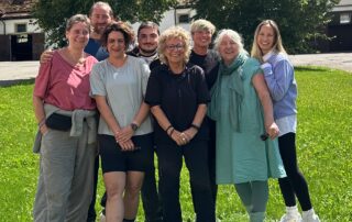 Beate Müller-Gemmeke steht lachend in der Mitte einer Gruppe von acht Personen auf einer Wiese vor einem landwirtschaftlichen Gebäude. Alle lächeln, es herrscht eine entspannte, herzliche Stimmung. Es sind alles ehemalige Mitarbeiter:innen von Beate.