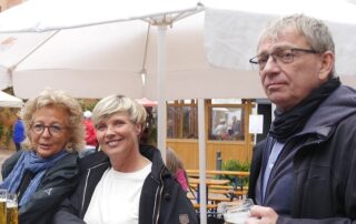 Beate Müller-Gemmeke steht mit Cindy Holmberg (MdL) und Martin draußen an einem Tisch unter weißen Sonnenschirmen. Alle drei trinken etwas und wirken in ein Gespräch vertieft.