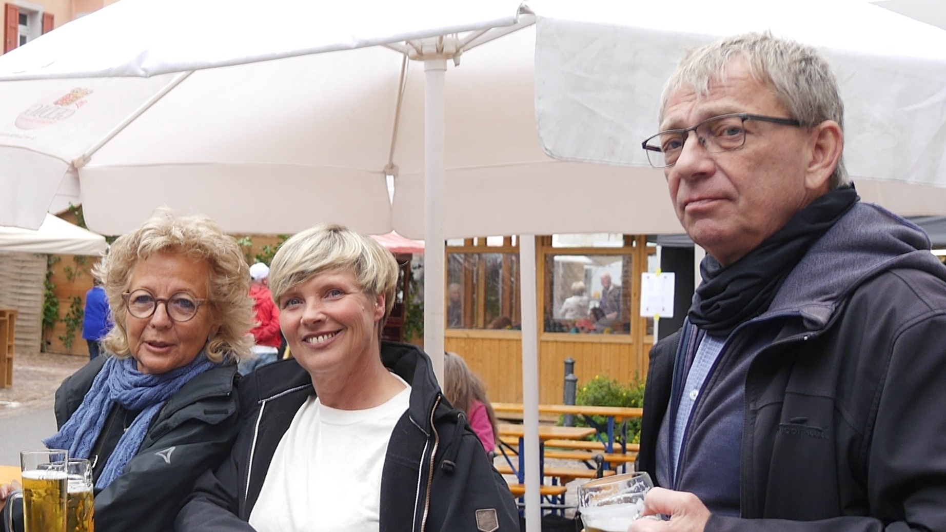 Beate Müller-Gemmeke steht mit Cindy Holmberg (MdL) und Martin draußen an einem Tisch unter weißen Sonnenschirmen. Alle drei trinken etwas und wirken in ein Gespräch vertieft.