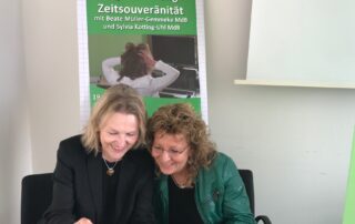 Beate Müller-Gemmeke und Sylvia Kotting-Uhl sitzen nebeneinander vor einem Plakat zur Digitalisierung, beide schauen lachend auf ein Handy, weil sie ein Selfie machen wollen.