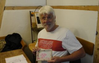Florian sitzt in einem Gastraum an einem Holztisch und hält ein kleines Bilderrahmen-Schild mit der Aufschrift „Müller“. Er trägt ein T-Shirt mit dem Aufdruck „Schwaben Power“ und lächelt in die Kamera.