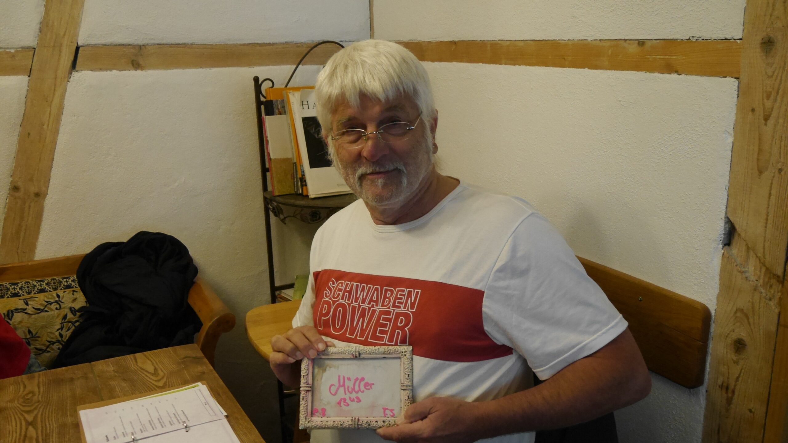 Florian sitzt in einem Gastraum an einem Holztisch und hält ein kleines Bilderrahmen-Schild mit der Aufschrift „Müller“. Er trägt ein T-Shirt mit dem Aufdruck „Schwaben Power“ und lächelt in die Kamera.