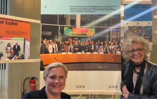 Beate Müller-Gemmeke und Steffi Aeffner bei einer Ausstellung, im Hintergrund ein großes Gruppenfoto des Gewerkschafts- und Sozialbeirats.