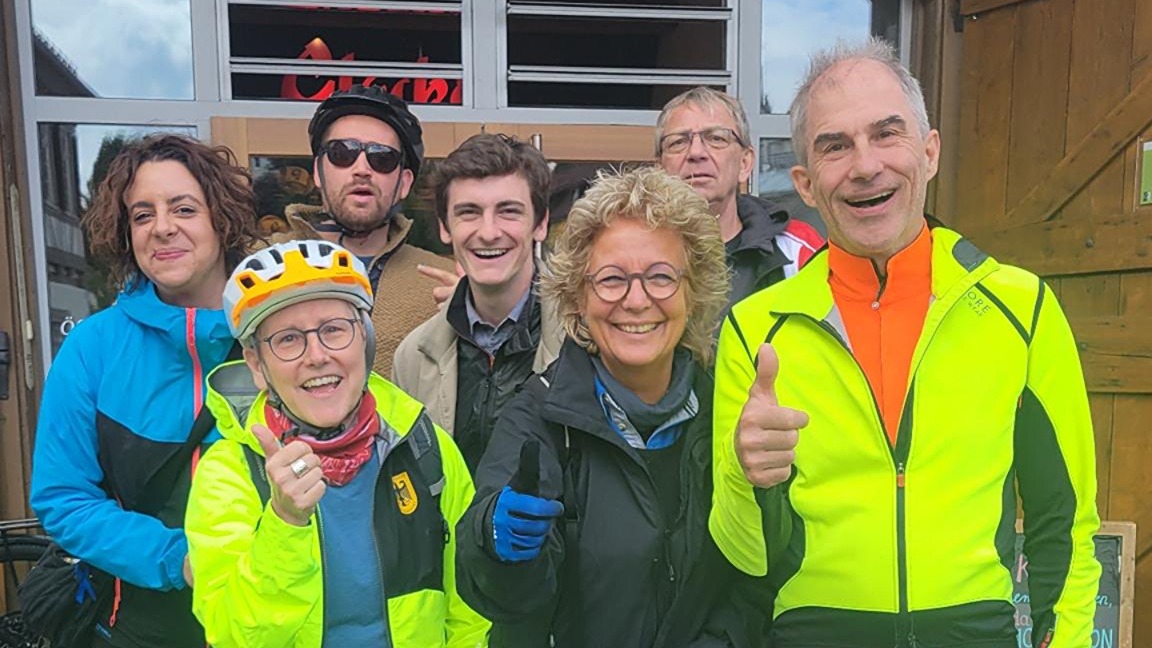 Gruppenbild vor einem Gasthaus mit Radfahrer:innen und Begleiter:innen. In der Mitte Beate Müller-Gemmeke, rechts und links das Albtour-Team, einige mit Fahrradhelmen und Jacken. Alle lachen oder zeigen Daumen hoch.