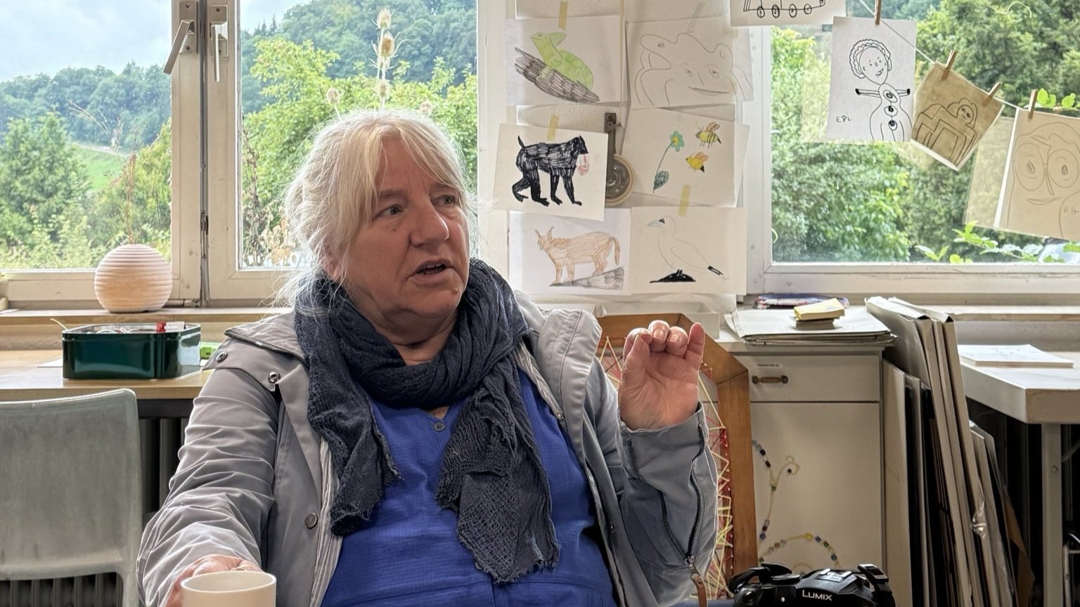 Hanne sitzt mit einer Tasse in der Hand in einem Raum mit vielen Zeichnungen an der Wand. Sie spricht gerade und wirkt konzentriert und engagiert.