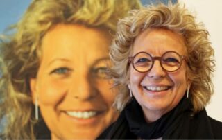 Beate Müller-Gemmeke vor einem großen Porträt von sich selbst (vor 16 Jahren), sie lächelt direkt in die Kamera.