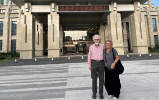Beate Müller-Gemmeke steht gemeinsam mit Prof. Dr. Wolfgang Däubler vor dem Haupteingang eines großen, modernen Gebäudes in Chengdu. Über dem Eingang hängt eine rote LED-Anzeigetafel mit chinesischer Begrüßung. Beide lächeln in die Kamera, sonniges Wetter.