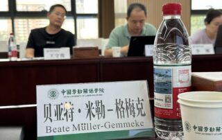 Namensschild von Beate Müller-Gemmeke auf einem Konferenztisch an der China University of Labour Relations in Peking, daneben eine Wasserflasche und ein Pappbecher mit Uni-Logo, im Hintergrund Teilnehmende bei der Arbeit.