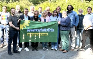 Beate Müller-Gemmeke mit Teilnehmer:innen der Jahrestagung von GewerkschaftsGrün. Die Gruppe hält ein grünes Banner mit Sonnenblume und dem Schriftzug „Gerechte Löhne & soziale Sicherheit“.