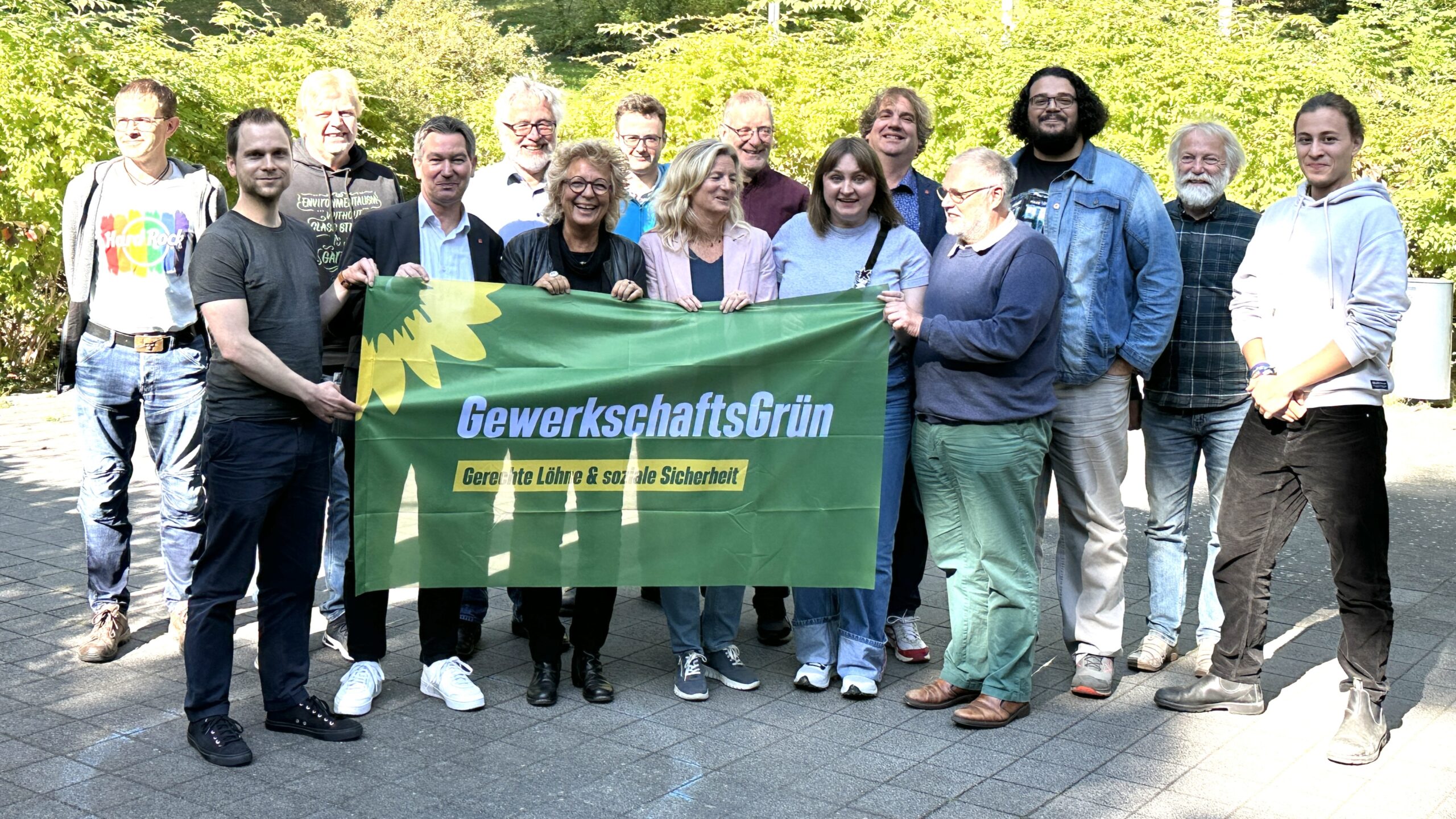 Beate Müller-Gemmeke mit Teilnehmer:innen der Jahrestagung von GewerkschaftsGrün. Die Gruppe hält ein grünes Banner mit Sonnenblume und dem Schriftzug „Gerechte Löhne & soziale Sicherheit“.