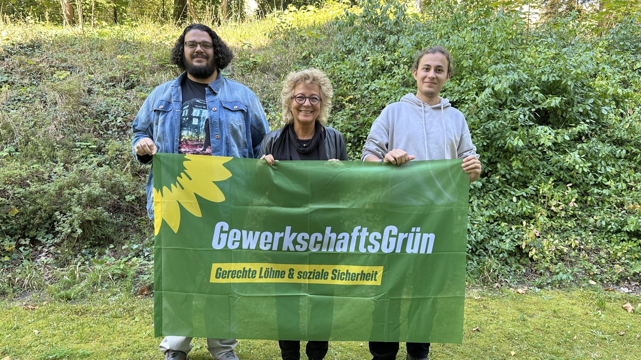 Beate Müller-Gemmeke mit zwei baden-württembergischen Mitgliedern von GewerkschaftsGrün Baden-Württemberg. Sie halten ein Banner mit Sonnenblume und dem Schriftzug „Gerechte Löhne & soziale Sicherheit“.