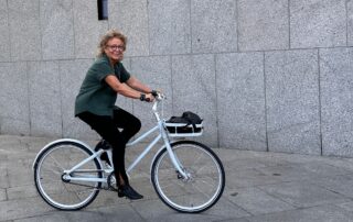 Beate Müller-Gemmeke fährt lachend mit einem weißen Fahrrad an einem grauen Steinbau entlang. Sie trägt eine grüne Bluse, schwarze Hose und schwarze Schuhe.
