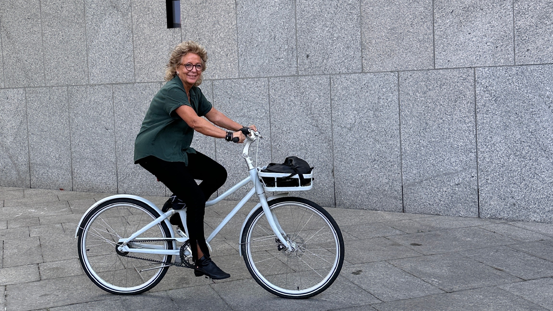 Beate Müller-Gemmeke fährt lachend mit einem weißen Fahrrad an einem grauen Steinbau entlang. Sie trägt eine grüne Bluse, schwarze Hose und schwarze Schuhe.
