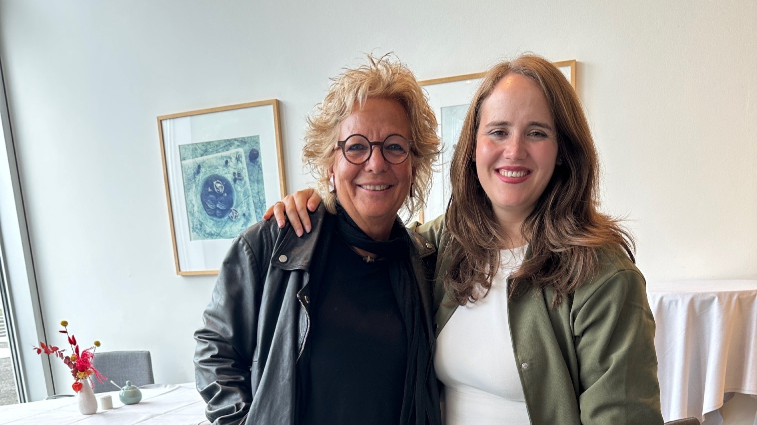 Beate Müller-Gemmeke und Ricarda Lang stehen nebeneinander, lachen in die Kamera und haben die Arme umeinander gelegt. Beate trägt eine schwarze Lederjacke, Ricarda ein weißes Kleid mit olivgrüner Jacke.