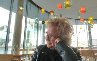 Beate Müller-Gemmeke sitzt in einem modernen Café mit bunten Lampen, stützt den Kopf auf die Hand und schaut nachdenklich zur Seite. Sie trägt eine schwarze Lederjacke und Brille.
