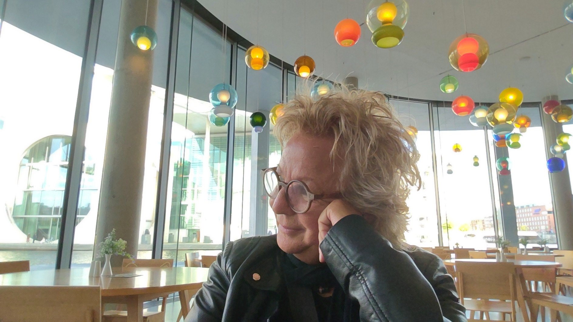 Beate Müller-Gemmeke sitzt in einem modernen Café mit bunten Lampen, stützt den Kopf auf die Hand und schaut nachdenklich zur Seite. Sie trägt eine schwarze Lederjacke und Brille.