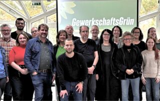 Beate Müller-Gemmeke mit Teilnehmer:innen der Jahrestagung von GewerkschaftsGrün. Alle stehen gemeinsam vor einer Leinwand mit dem Logo von GewerkschaftsGrün.