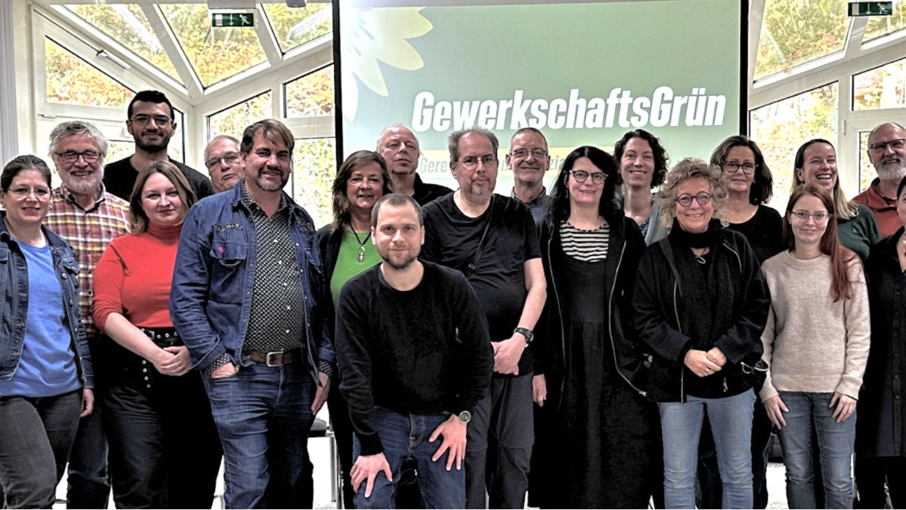 Beate Müller-Gemmeke mit Teilnehmer:innen der Jahrestagung von GewerkschaftsGrün. Alle stehen gemeinsam vor einer Leinwand mit dem Logo von GewerkschaftsGrün.