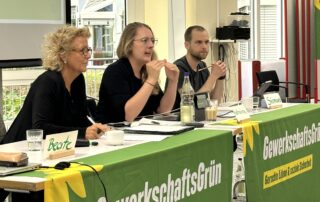 Beate Müller-Gemmeke mit der Fraktionsvorsitzenden der grünen Bundestagsfraktion – Katharina Dröge, die Gast bei der Jahrestagung von GewerkschaftsGrün ist. Daneben ist Simon Dylla, der Co-Bundessprecher von GewerkschaftsGrün. Sie sitzen am Podium vor einem grünen Banner mit dem Logo von GewerkschaftsGrün.