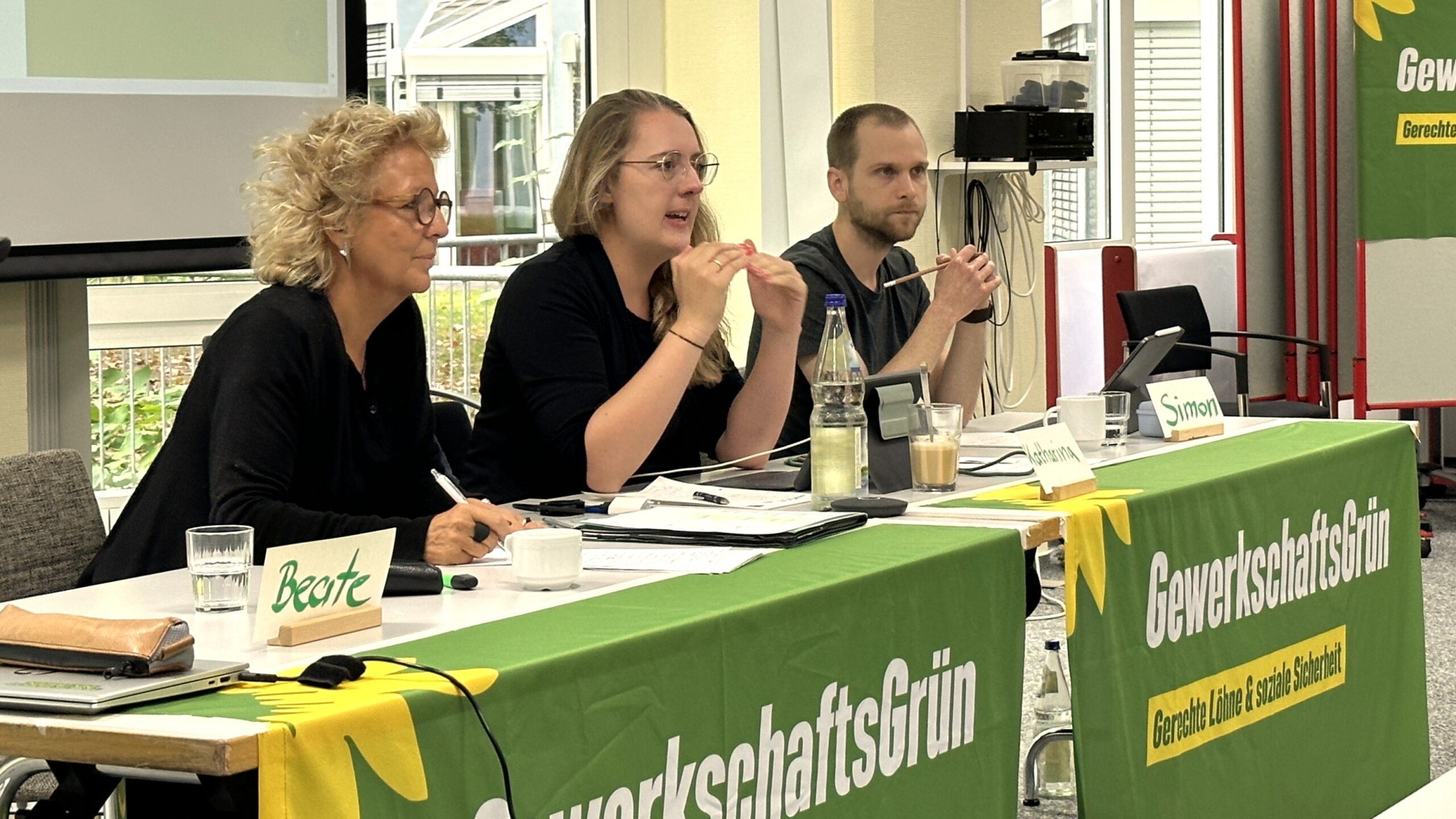 Beate Müller-Gemmeke mit der Fraktionsvorsitzenden der grünen Bundestagsfraktion – Katharina Dröge, die Gast bei der Jahrestagung von GewerkschaftsGrün ist. Daneben ist Simon Dylla, der Co-Bundessprecher von GewerkschaftsGrün. Sie sitzen am Podium vor einem grünen Banner mit dem Logo von GewerkschaftsGrün.