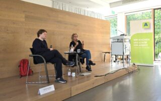 Beate Müller-Gemmeke sitzt mit einem Gesprächspartner auf einer kleinen Bühne, beide halten Mikrofone in der Hand. Im Hintergrund eine schlichte Holzverkleidung. Auf dem Tisch stehen Wasserflaschen und Tassen – eine ruhige Gesprächssituation im Rahmen der Arbeitslosentagung.
